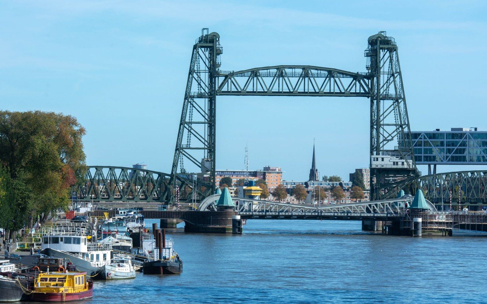 Steel lift bridge spans a river with moored boats and a city skyline behind