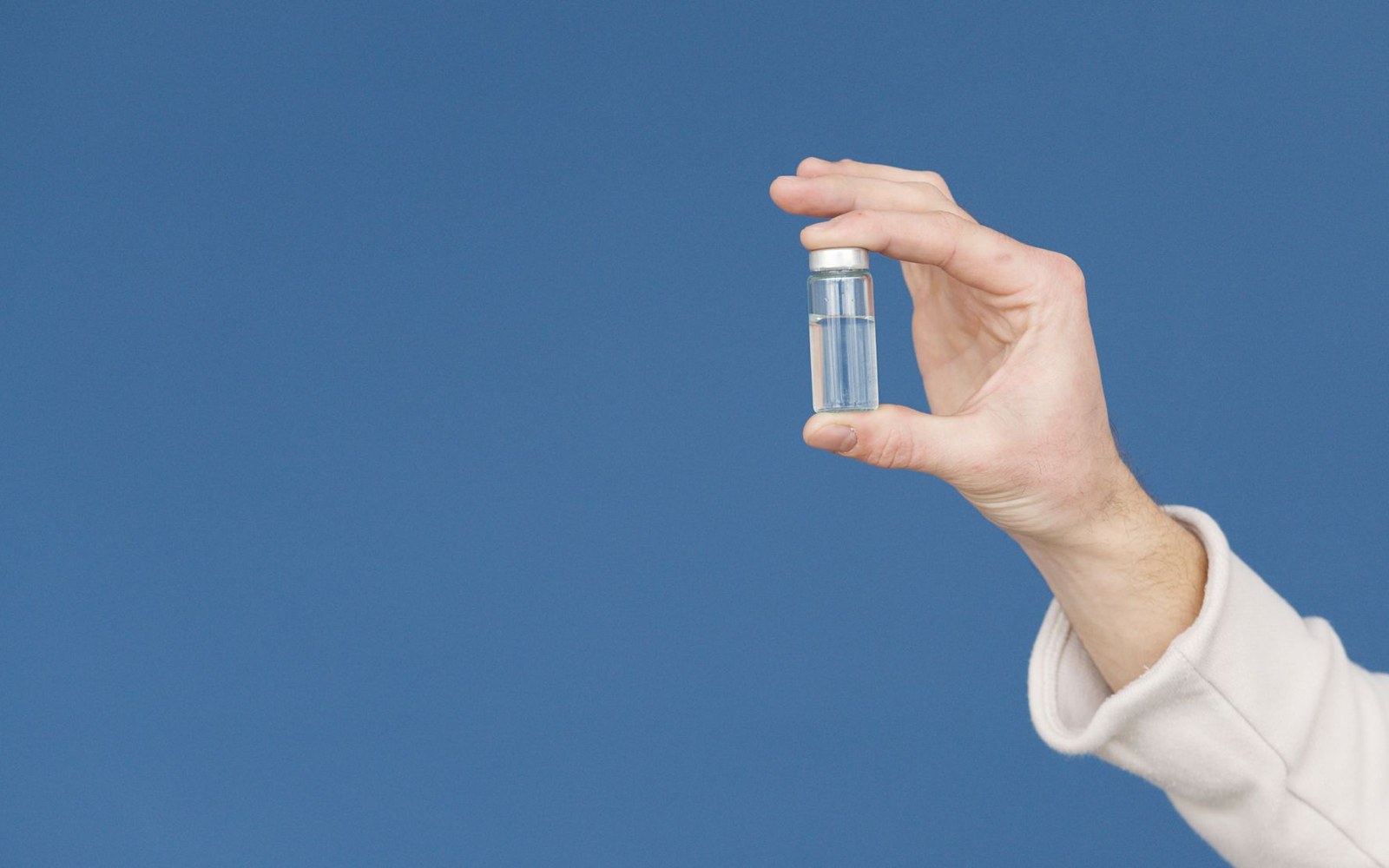 A hand holds a small glass vial containing clear liquid against a blue background
