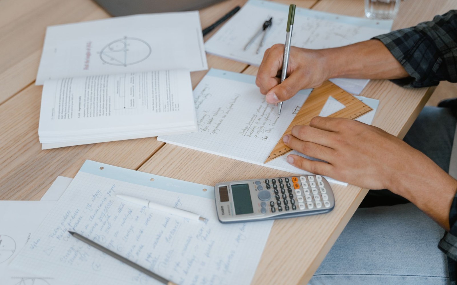 Hands writing math notes with a calculator, ruler, and compass on a wooden desk